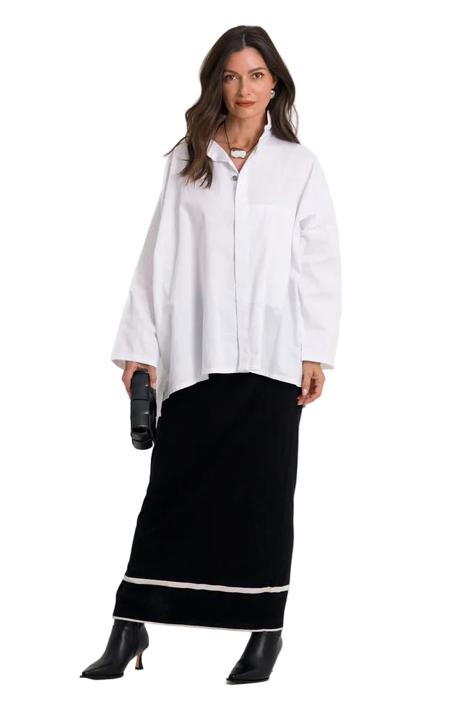 A woman modeling a white cotton long-sleeve button-up shirt paired with a black maxi skirt featuring a white horizontal stripe near the hem, and black pointed-toe heeled boots, against a plain white studio background.