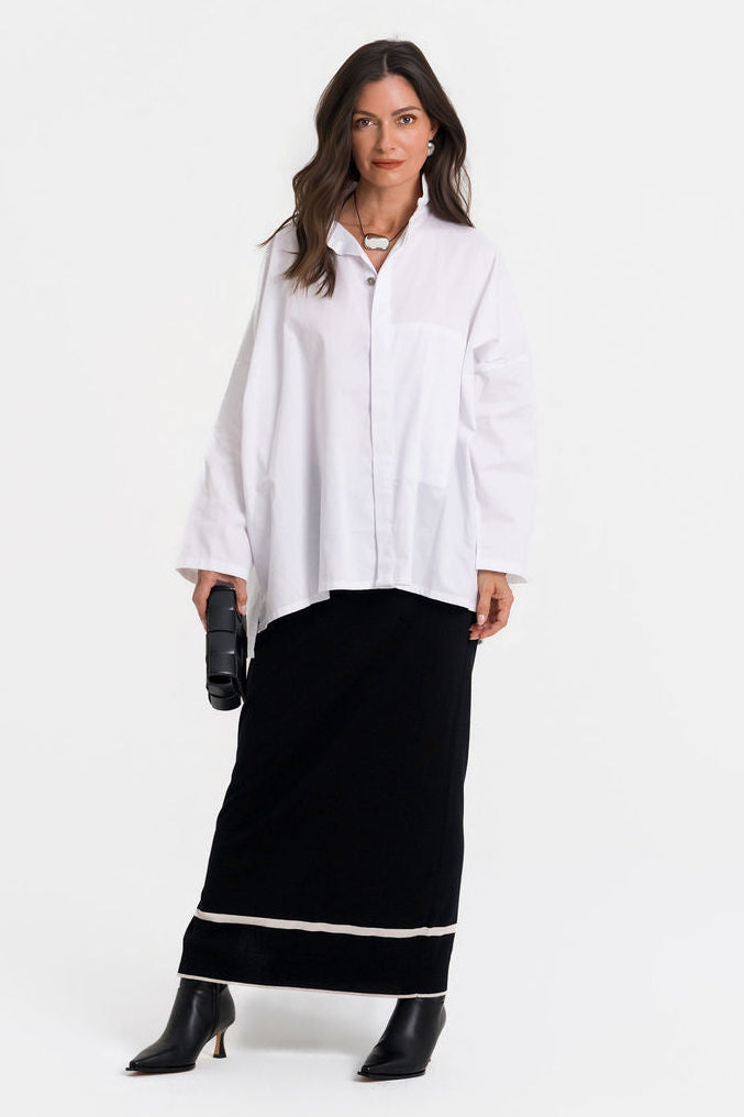 A woman modeling a white cotton long-sleeve button-up shirt paired with a black maxi skirt featuring a white horizontal stripe near the hem, and black pointed-toe heeled boots, against a plain white studio background.