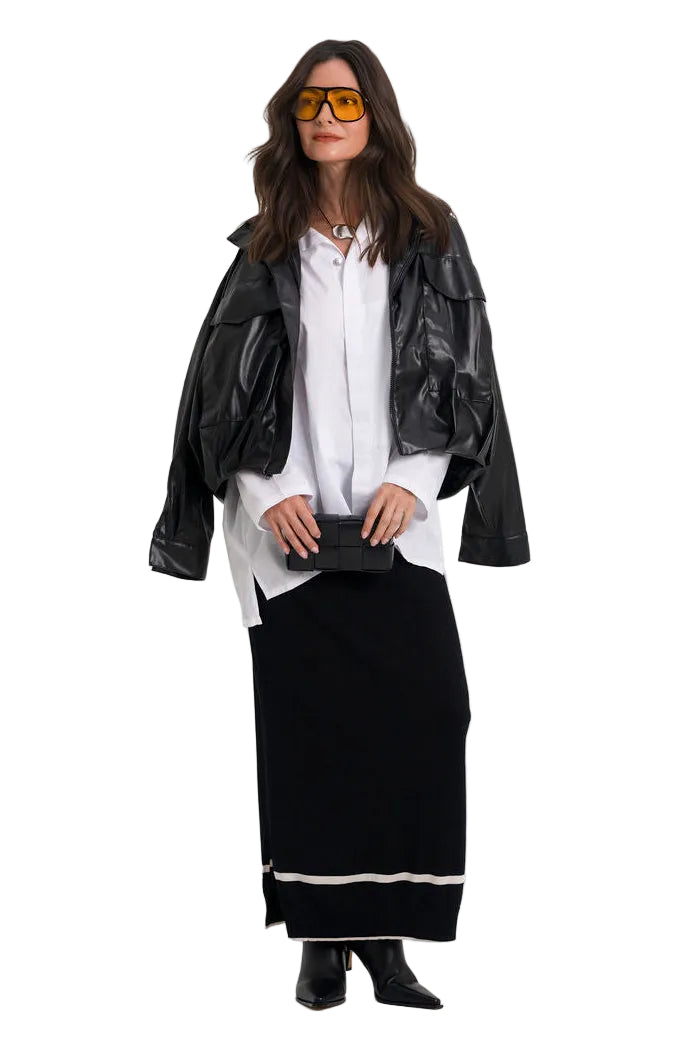 A woman modeling a black faux-leather moto jacket over a white button-down shirt and a long black skirt with a white stripe, styled with yellow-tinted sunglasses and a black clutch, on a solid white background.