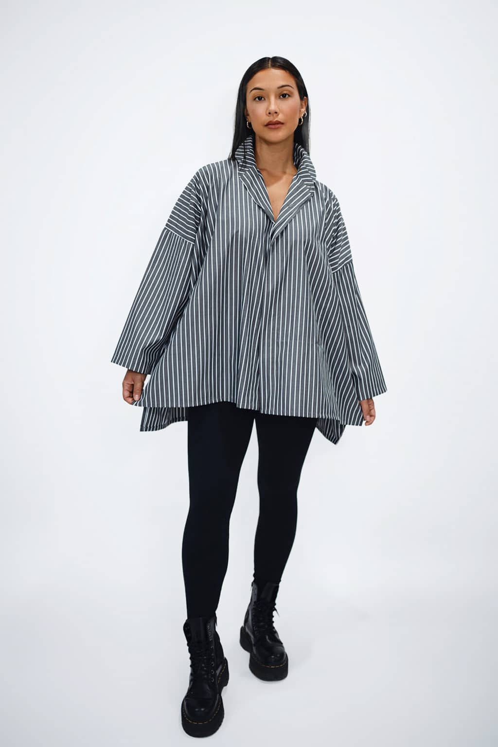 Woman wearing a black and white striped oversized Planet by Lauren G 100% cotton shirt with a popped collar, black leggings, and black boots on a white background