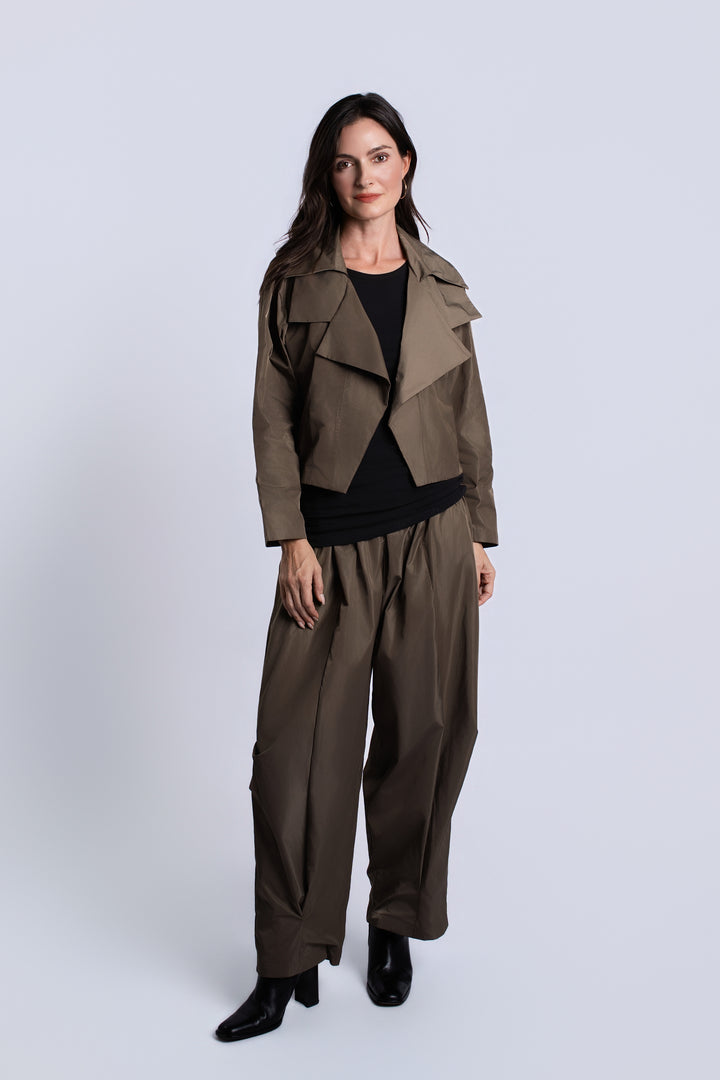 A woman modeling a coordinated outfit consisting of a cropped taupe-brown jacket, wide-leg taupe-brown trousers, a black top, and black ankle boots, photographed against a light gray studio background.