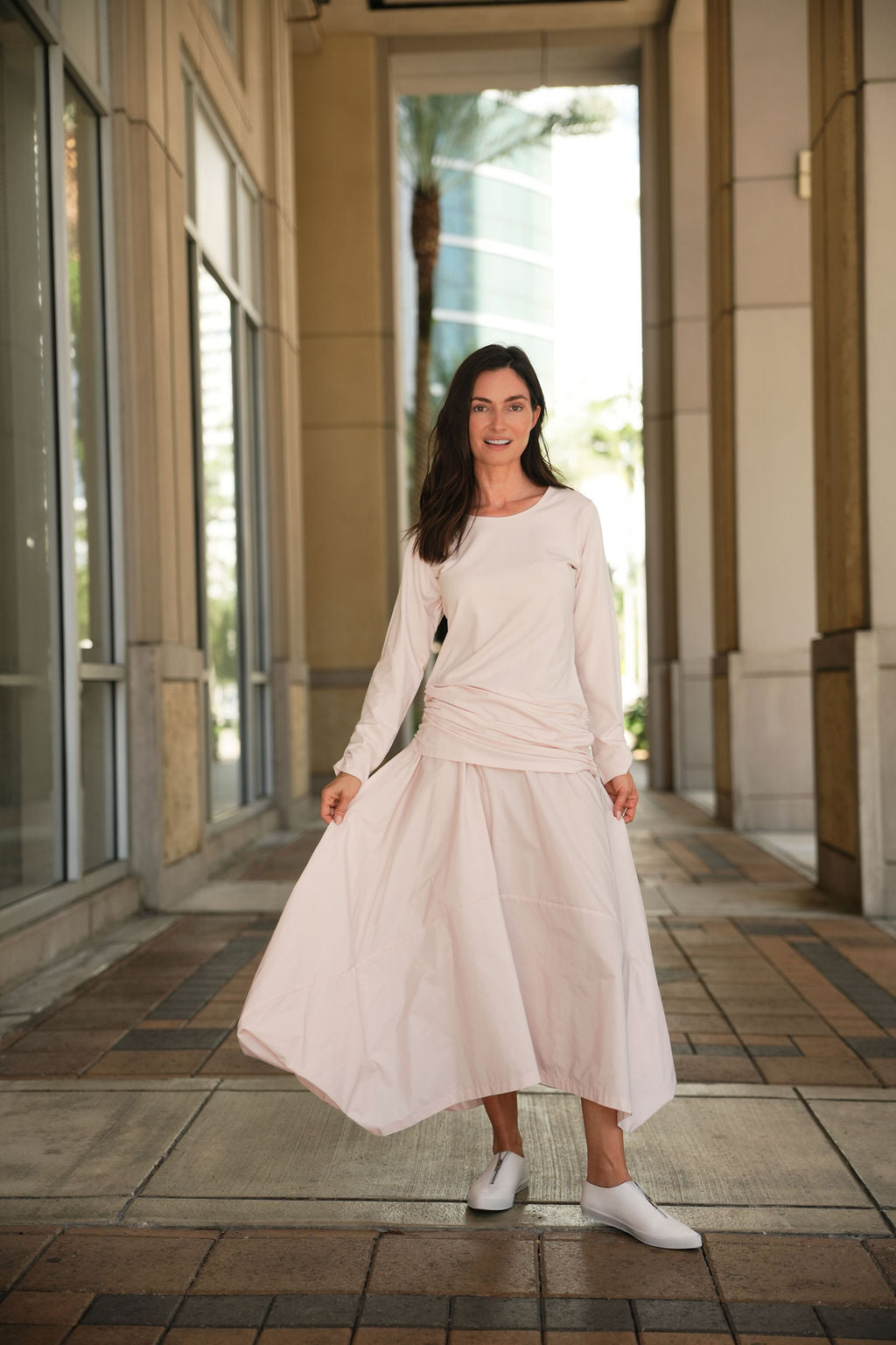 Woman in a blush skirt and cotton lycra ruche top standing in an outdoor setting with large windows in the background