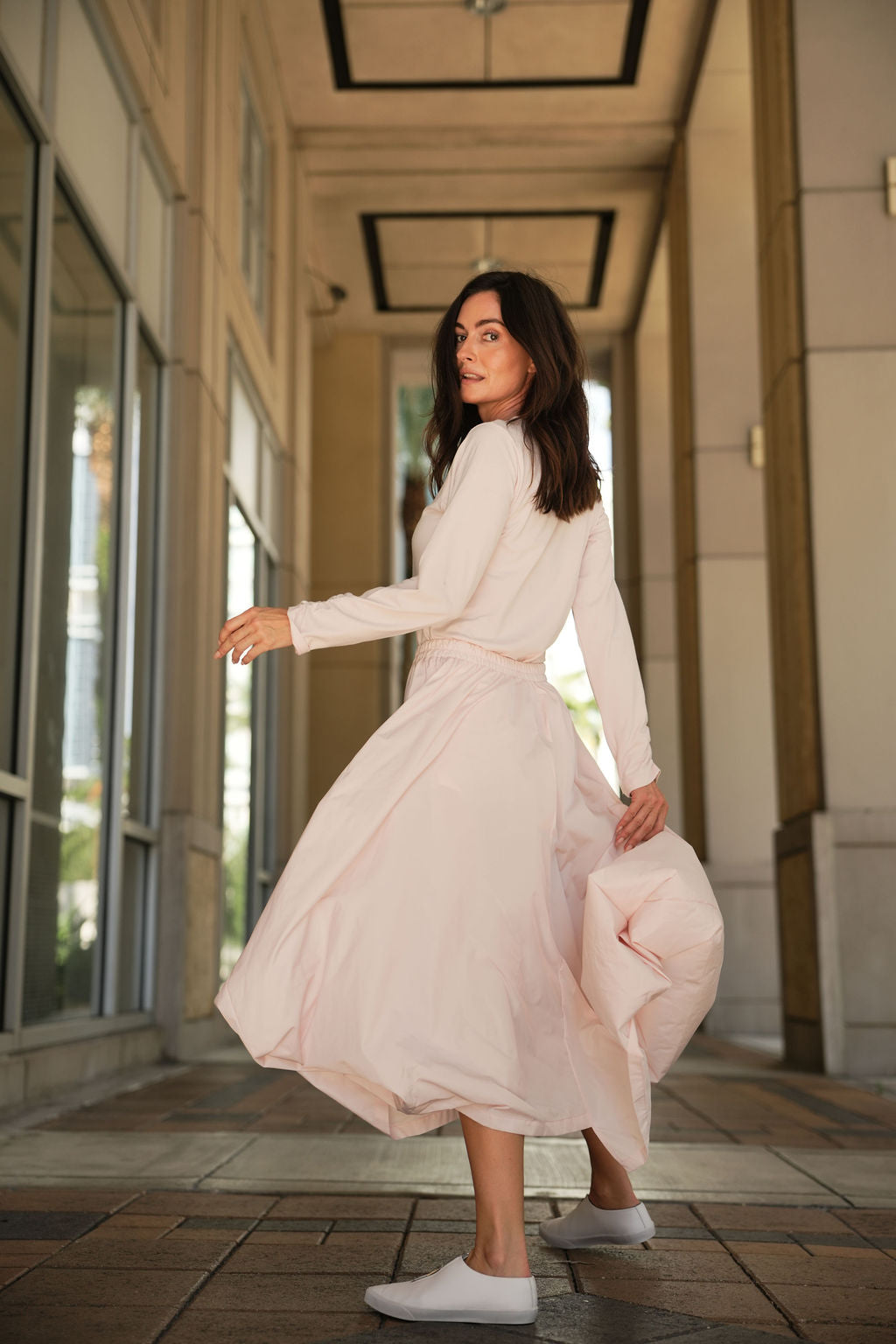 Woman in a pink blush skirt standing in an outdoor setting near large windows.