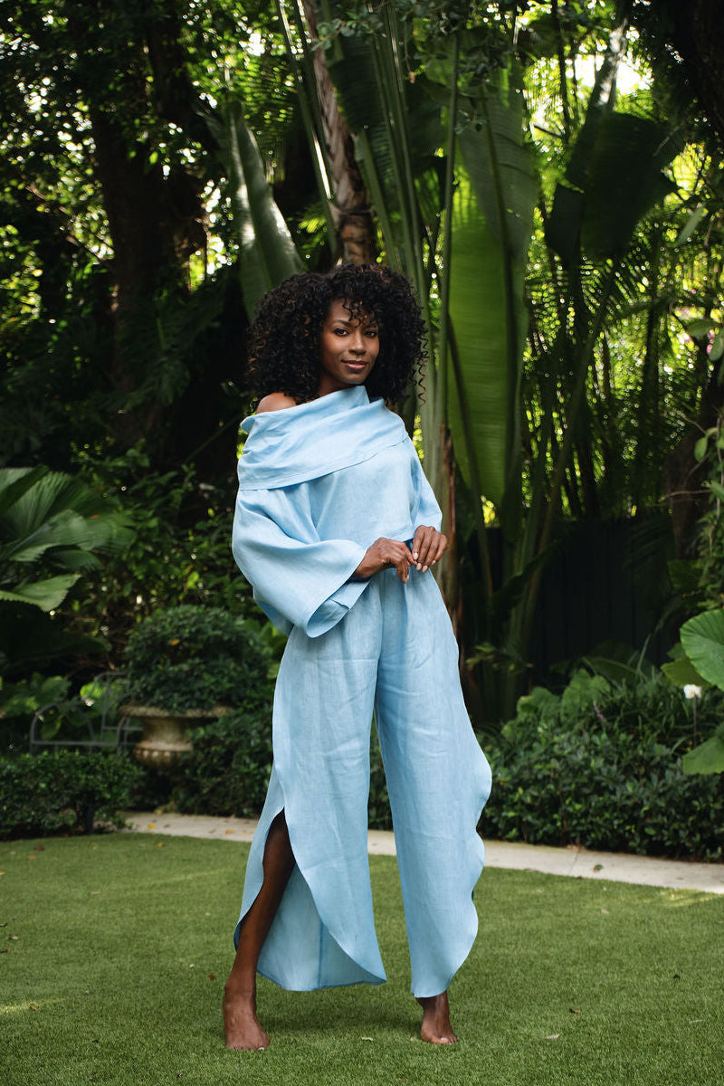 A woman stands barefoot on a green lawn in a lush garden, wearing a 100% linen flowing light blue draped garment with wide sleeves and a high slit.