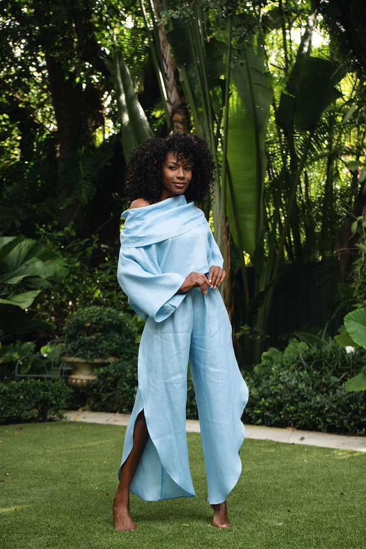 A woman stands barefoot on a green lawn in a lush garden, wearing a 100% linen flowing light blue draped garment with wide sleeves and a high slit.