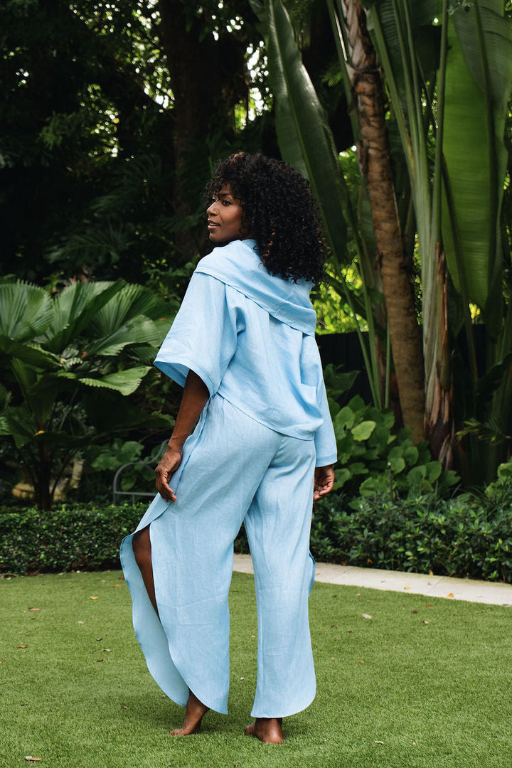 A woman stands barefoot on a green lawn in a lush garden, wearing a light blue matching lounge set consisting of a 100% linen short-sleeve top and wide-leg pants.