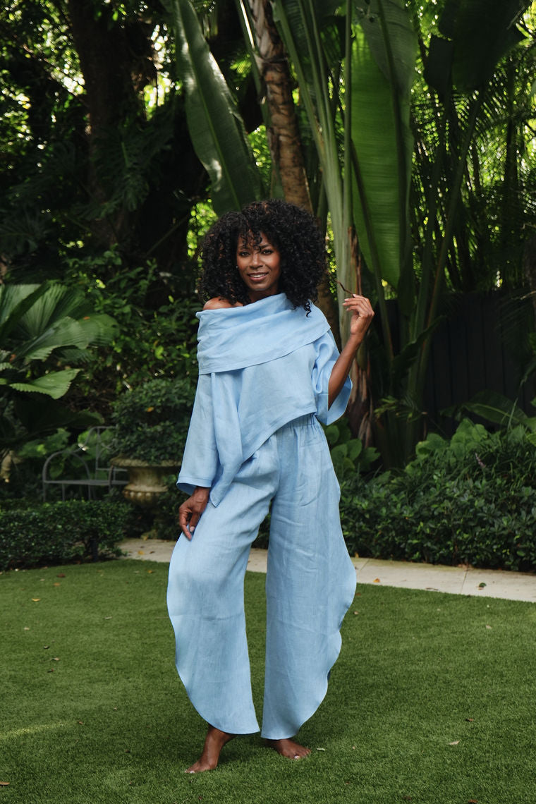 A woman stands barefoot on green grass in a lush garden, wearing a light blue 100% linen off-the-shoulder draped jumpsuit or matching set with wide-leg pants.