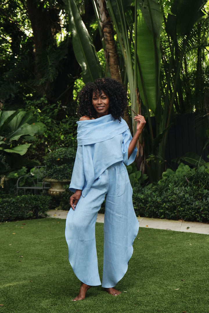 A woman stands barefoot on green grass in a lush garden, wearing a light blue 100% linen off-the-shoulder draped jumpsuit or matching set with wide-leg pants.