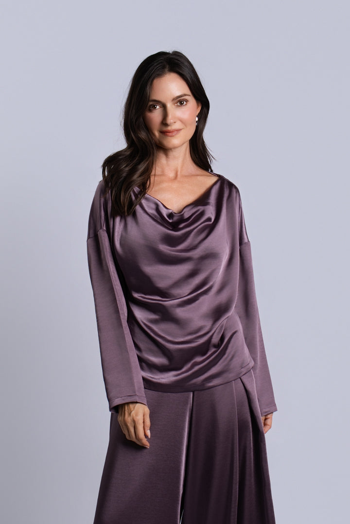 A woman modeling a matching two-piece satin outfit in a dusty plum purple color, featuring a draped long-sleeve top and wide-leg trousers, set against a solid light gray studio background.