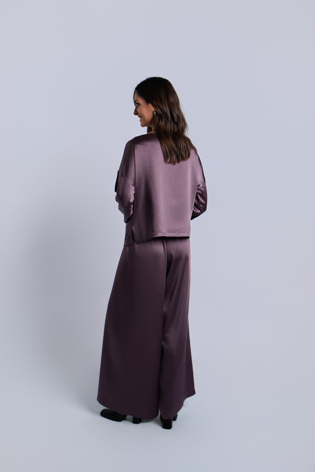 A woman is shown from the back wearing a matching amethyst purple satin two-piece set consisting of a cropped long-sleeve top and wide-leg trousers, set against a plain light gray studio background.