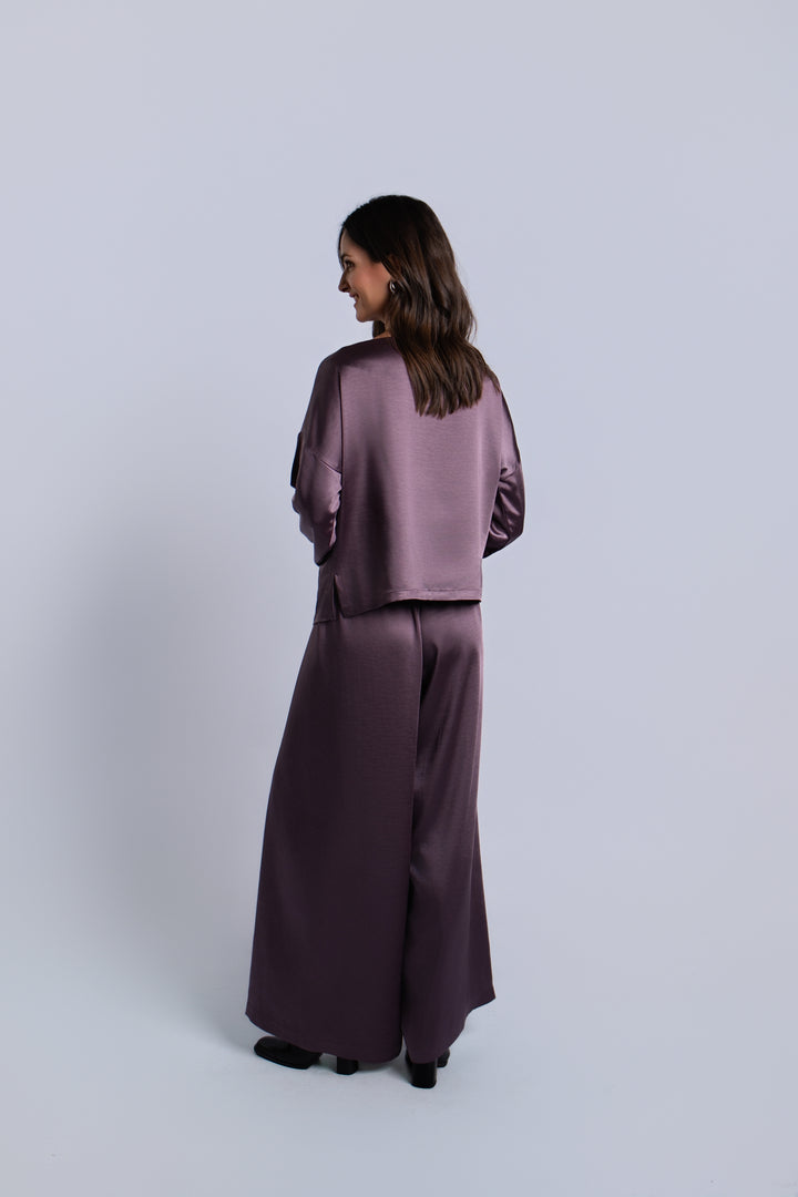 A woman is shown from the back wearing a matching amethyst purple satin two-piece set consisting of a cropped long-sleeve top and wide-leg trousers, set against a plain light gray studio background.
