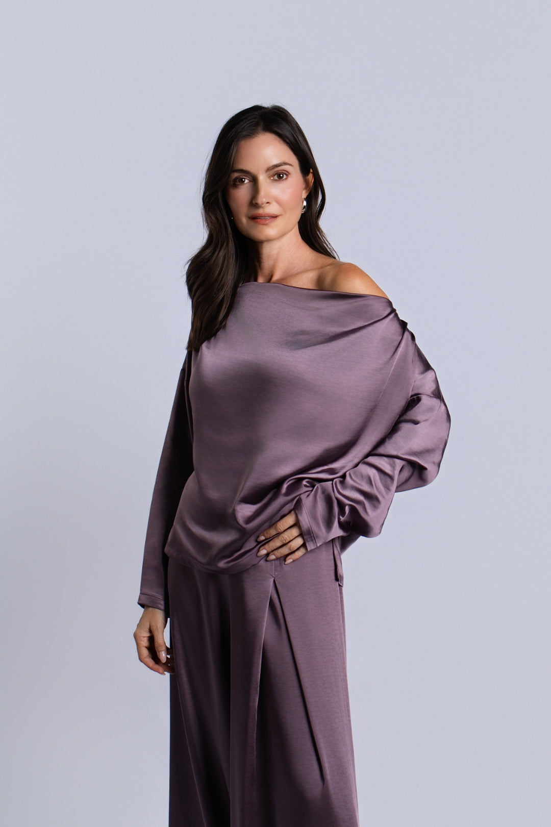 A woman modeling a matching off-the-shoulder long-sleeve sateen top and wide-leg trousers set in an amethyst purple satin fabric, set against a solid light gray studio background.