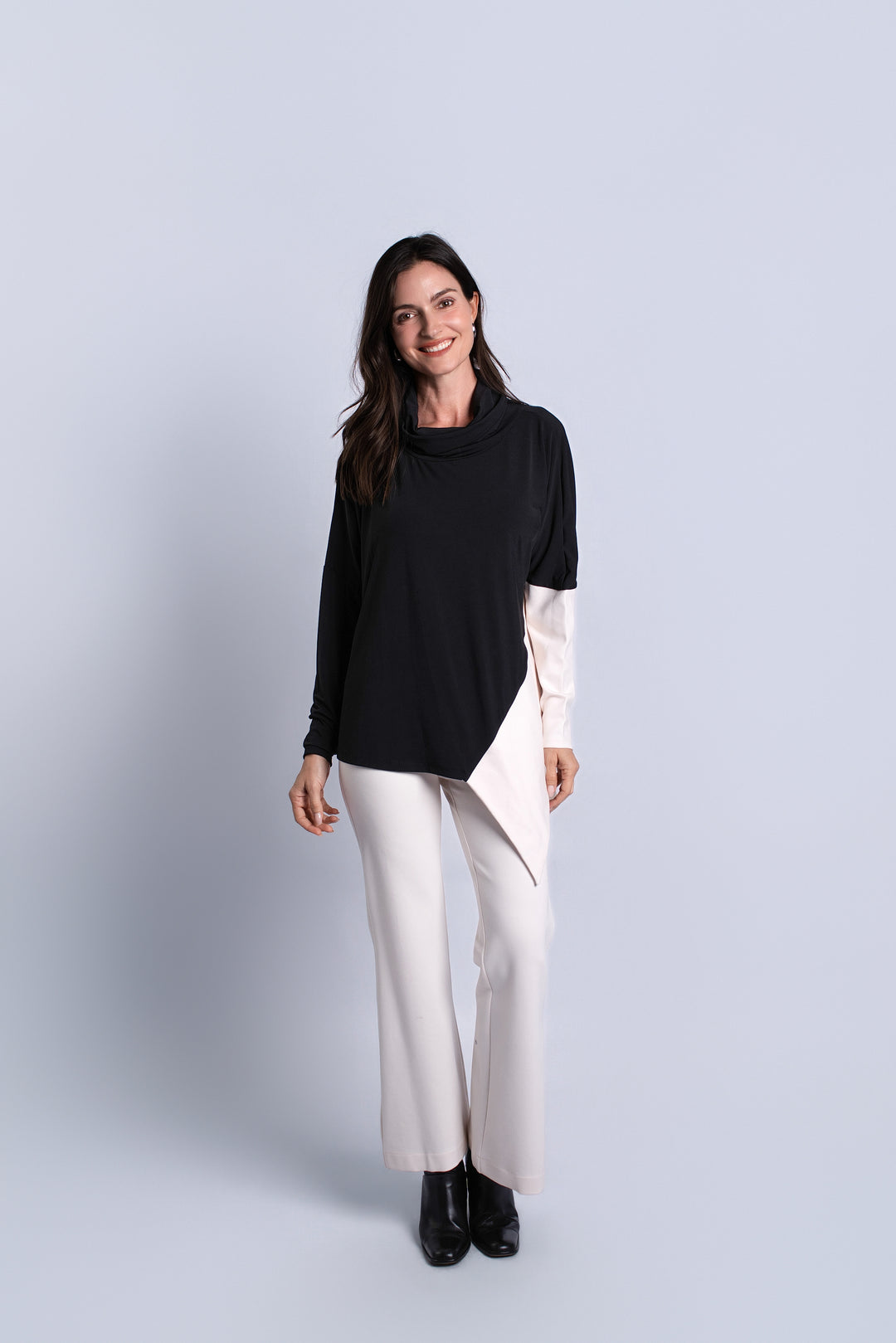 A woman modeling a color-blocked long-sleeve top with a black upper section and a light cream lower section, paired with cream wide-leg trousers and black ankle boots, against a light gray studio background.