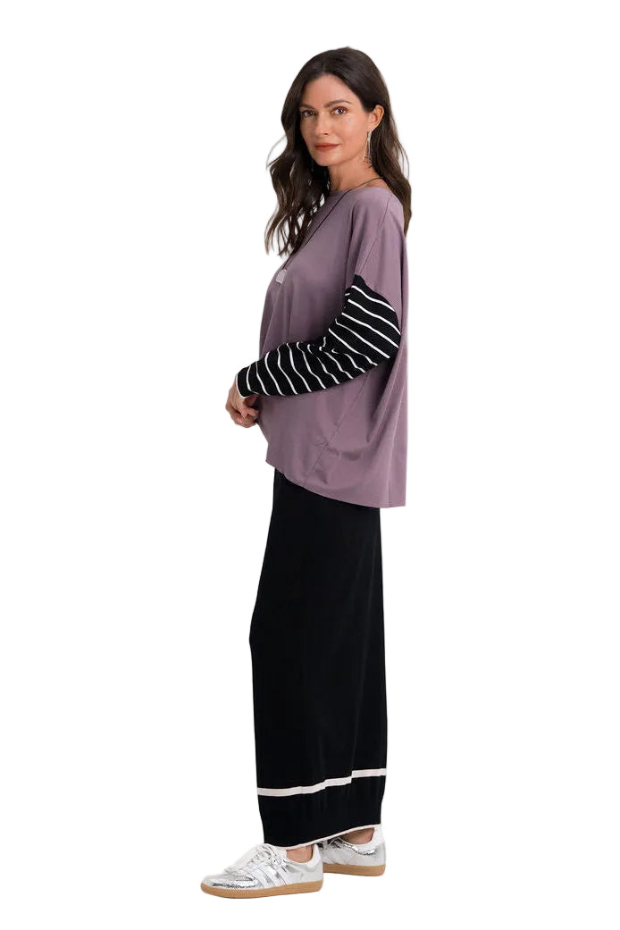 A woman modeling a layered outfit consisting of a 100% pima cotton lavender-purple long-sleeve top with black and white striped sleeves, paired with black wide-leg pants featuring white stripes at the hem, and white sneakers, against a plain white studio background.