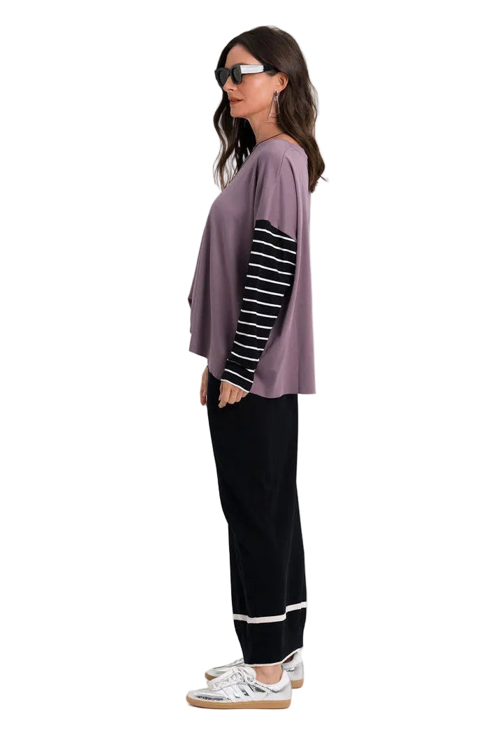 A woman stands in profile wearing a loose 100% pima cotton mauve-purple top with black-and-white striped sleeves, black pants, and white sneakers against a plain light gray studio background.