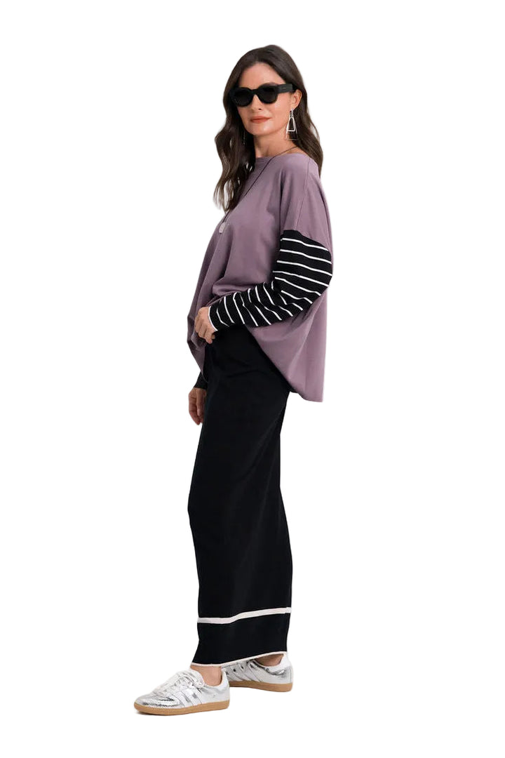 A woman modeling a 100% pima cotton lavender knit top with black and white striped sleeves, paired with black wide-leg pants and white sneakers, against a plain white studio background.