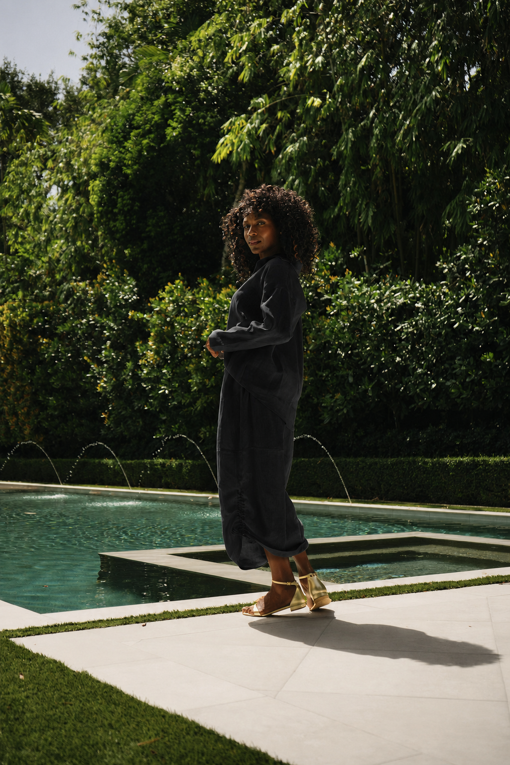 A woman with curly dark hair stands by a rectangular pool in a lush green outdoor setting, wearing a 100% linen shirt and skirt set and gold sandals.