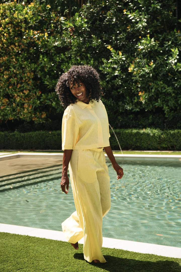 A woman with curly dark hair wearing a pale yellow short-sleeve collared top and matching 100% linen wide-leg trousers walks beside a swimming pool on a grassy lawn with a lush green hedge in the background.