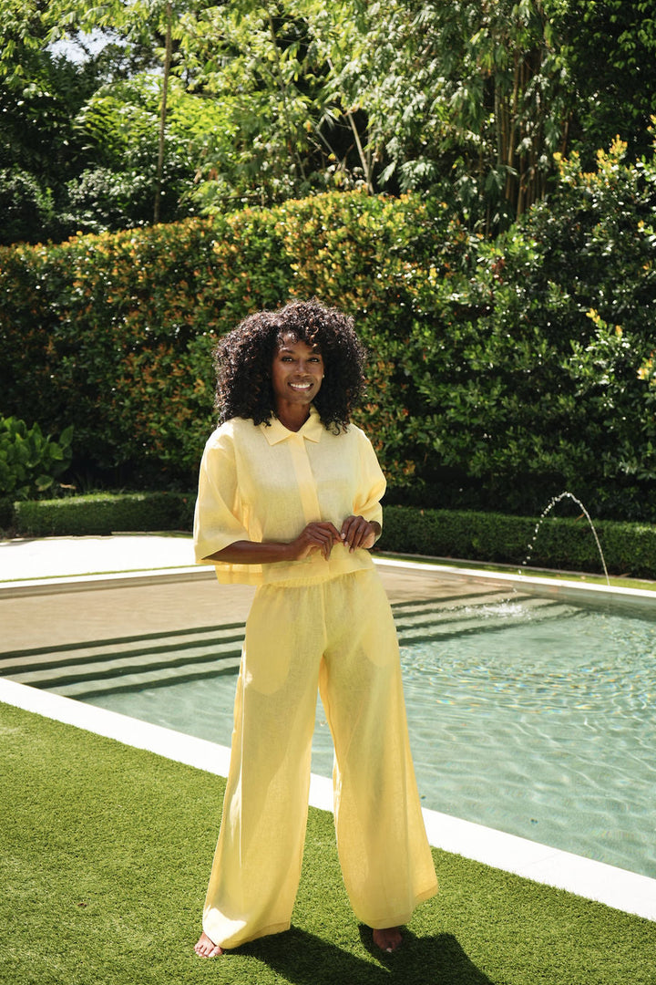 A woman with curly black hair stands outdoors beside a swimming pool on green grass, wearing a matching pale yellow collared top and 100% linen wide-leg pants set.