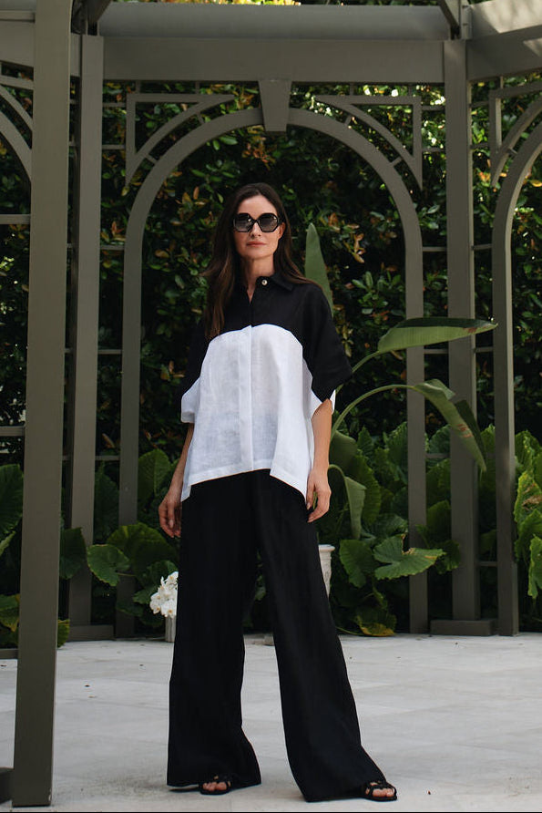 A woman wearing a black and white asymmetrical top and 100% linen black wide-leg pants stands under a large arched garden pavilion with green foliage in the background.