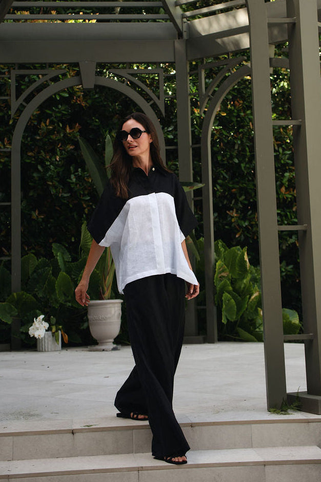 A woman wearing a black and white color-block short-sleeve top and 100% linen black wide-leg pants walks down stone steps under a large wooden pergola in a garden setting.