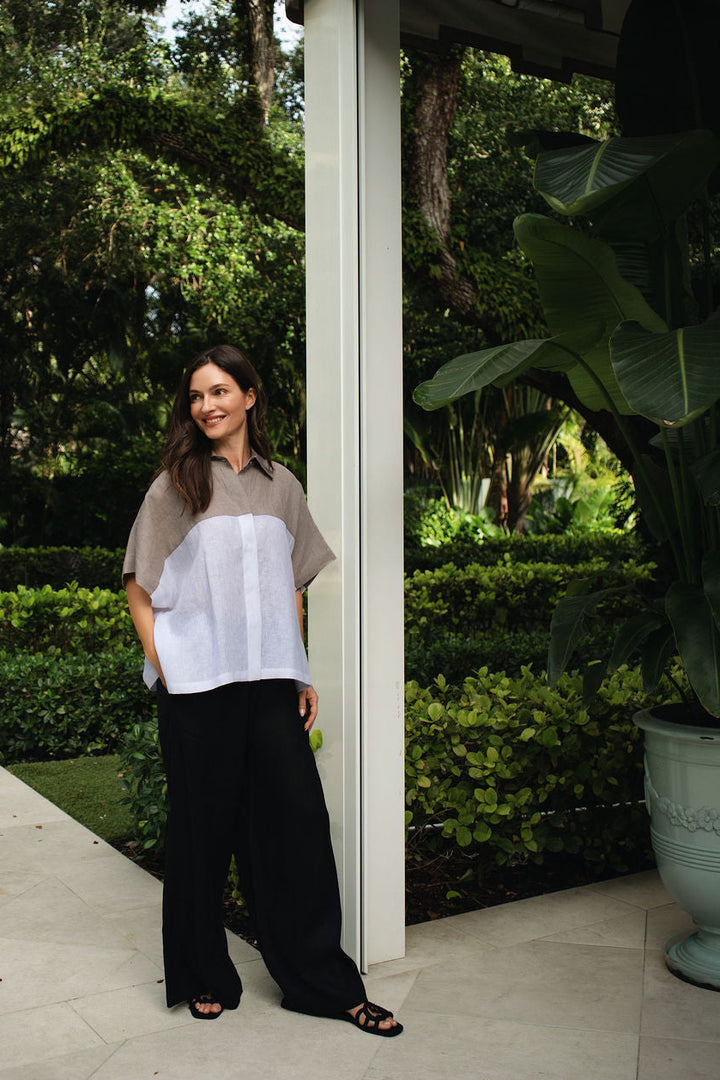 A woman stands on a patio beside a white column in a lush garden setting, wearing a short-sleeve collared top with a taupe and white color-block design, loose 100% linen black wide-leg pants, and black sandals.