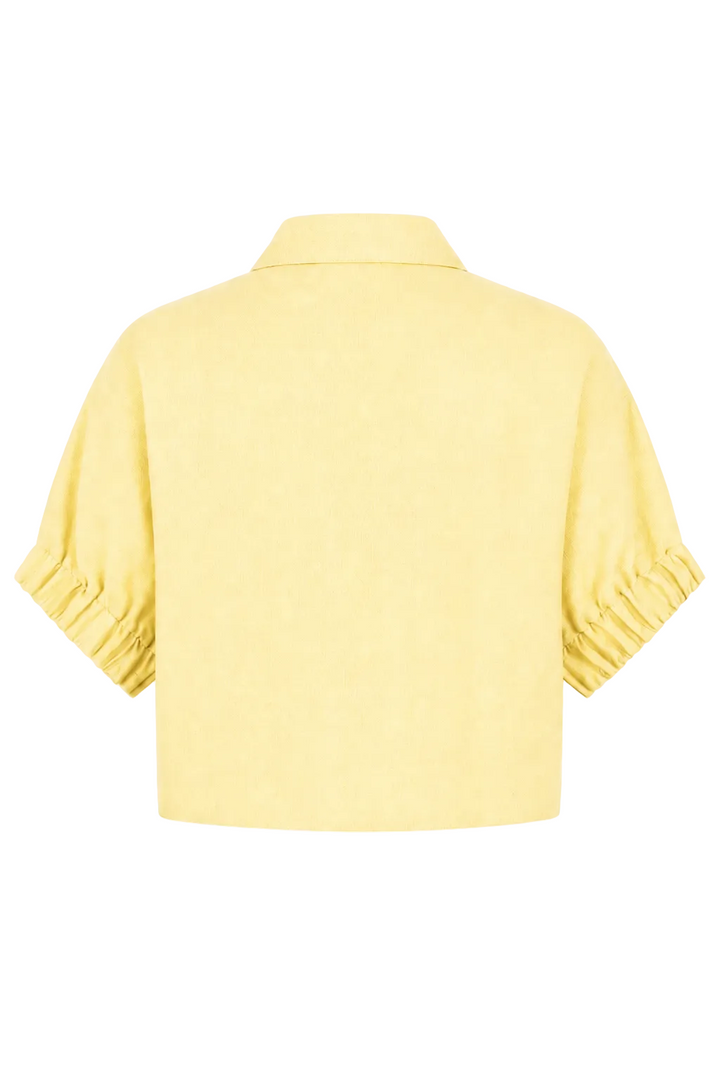 A 100% linen pale yellow short-sleeve collared top shown from the back, featuring gathered ruffle detailing at the cuffs, set against a plain white background.