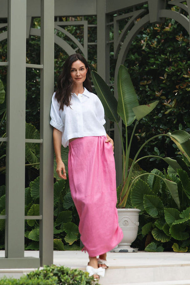 A woman stands on white steps under a large gray metal gazebo surrounded by lush green garden foliage. She is wearing a white short-sleeve crop top and a 100% linen bright pink wide-leg skirt.