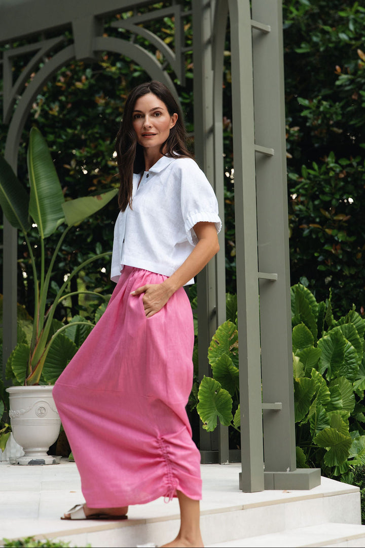 A woman posing outdoors in a garden setting under a gray metal trellis, wearing a white short-sleeve collared shirt and a bright pink 100% linen gathered maxi skirt.