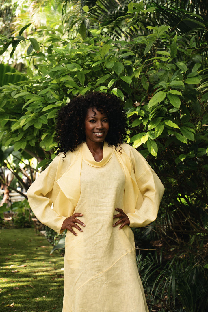 A woman with curly black hair wearing a banana yellow linen dress with voluminous long sleeves, standing in a lush green garden.