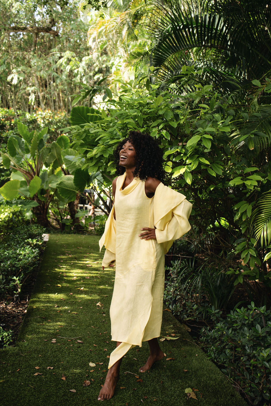 A woman wearing a banana yellow draped linen dress stands barefoot on a grassy garden path surrounded by lush tropical foliage.