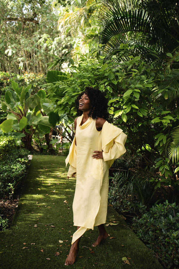 A woman wearing a banana yellow draped linen dress stands barefoot on a grassy garden path surrounded by lush tropical foliage.
