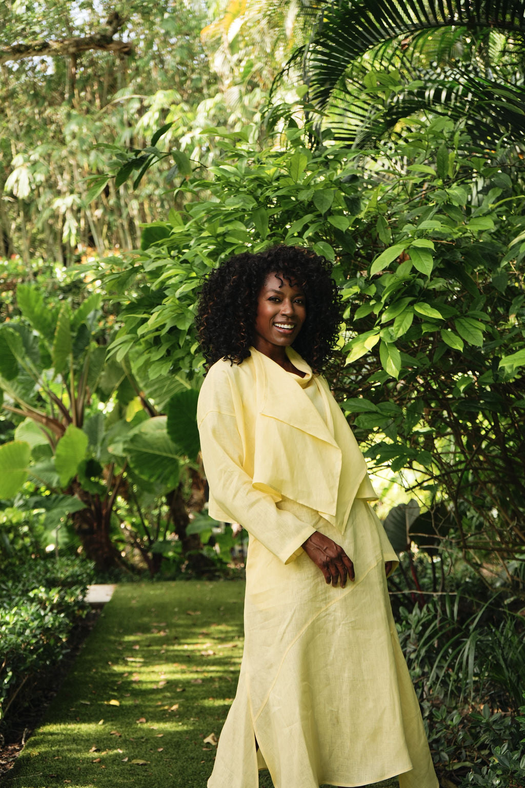 A woman with curly black hair wearing a banana yellow long-sleeve wrap dress, standing in a lush green tropical garden.