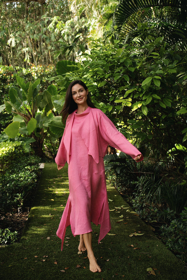 A woman wearing a matching fuchsia linen dress and lightweight draped jacket walks barefoot along a mossy garden path surrounded by lush green foliage.
