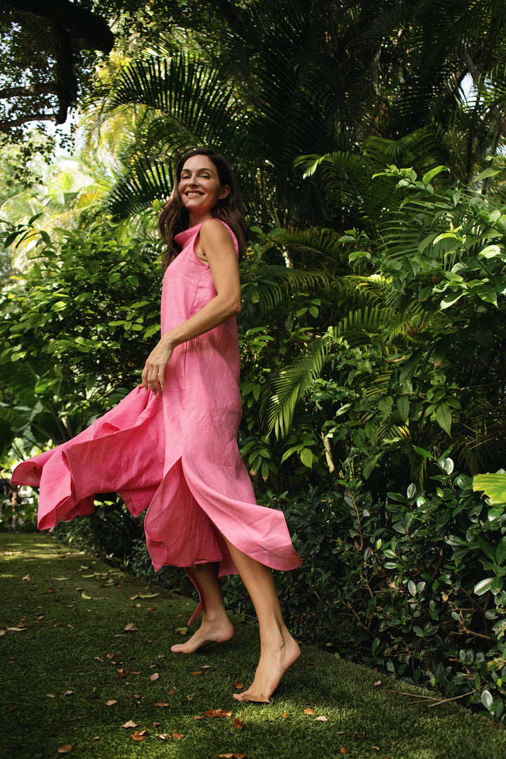 A woman wearing a flowing pink sleeveless linen dress stands barefoot in a lush green garden, with the dress blowing in the wind.