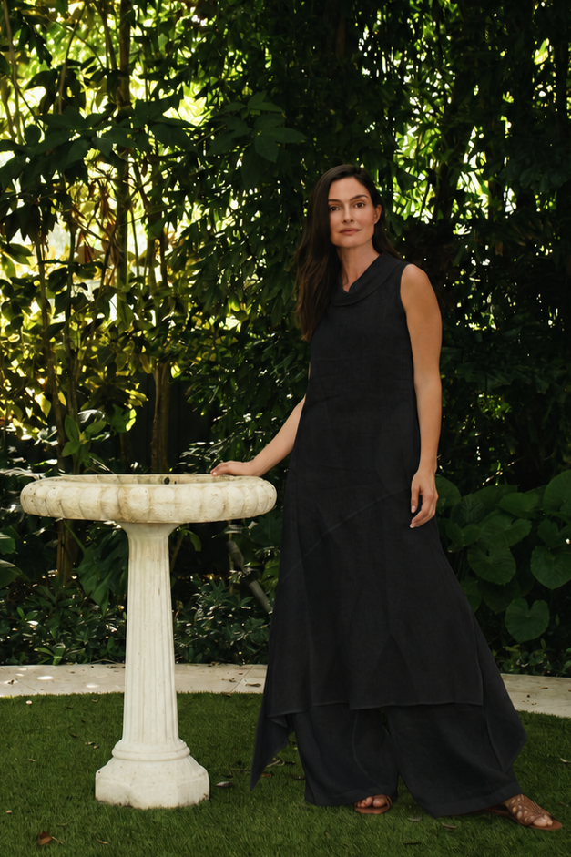 A woman in a 100% linen long black sleeveless dress stands beside a white stone birdbath in a lush garden with dense green foliage.