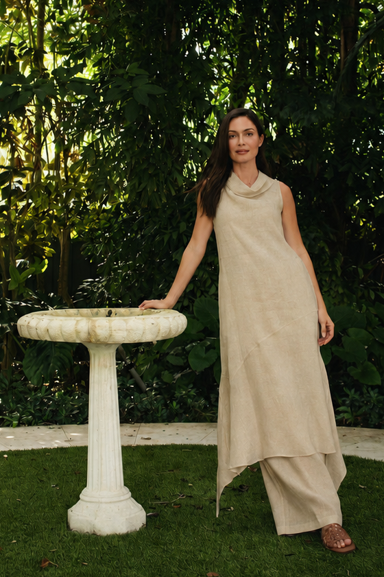 A woman wearing a beige 100% linen sleeveless maxi dress stands on a circular grassy lawn in a lush garden, touching a white stone birdbath.