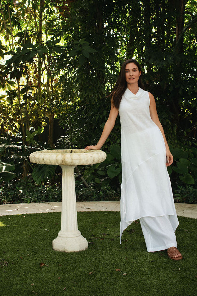 A woman wearing a white sleeveless linen dress stands on a grassy garden path next to a white stone birdbath, surrounded by a dense green forest background.