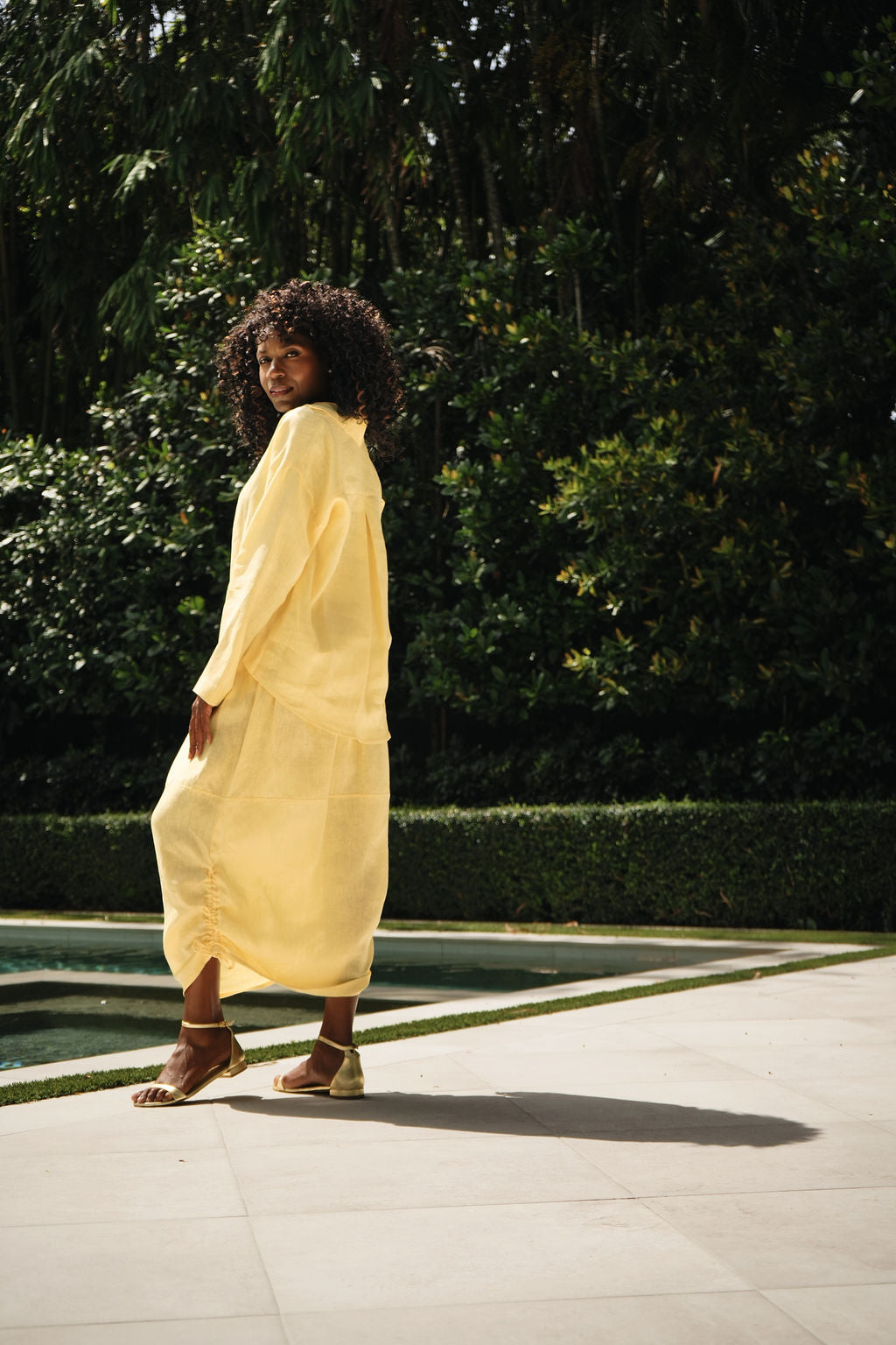 A woman wearing a flowing pale yellow 100% linen shirt and skirt set and brown strappy sandals walks along a paved path next to a pool with a lush green garden in the background.