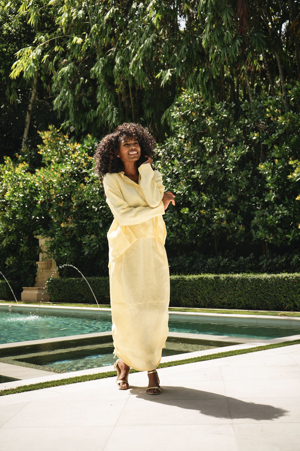 A woman stands outdoors by a pool wearing a long, loose, light yellow 100% linen shirt and skirt set or robe-style garment with brown sandals. Lush green trees and trimmed hedges are visible in the background.