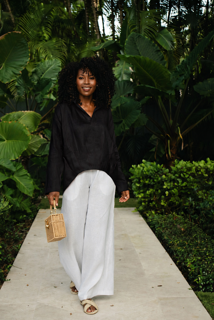 A woman walking on a stone path surrounded by lush tropical greenery, wearing a 100% linen black long-sleeve blouse, white wide-leg pants, and carrying a small woven tan handbag.