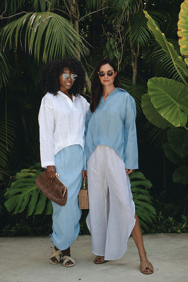 Two women standing outdoors in front of dense tropical green foliage, wearing 100% linen loose light blue and white outfits. One holds a brown leather bag.