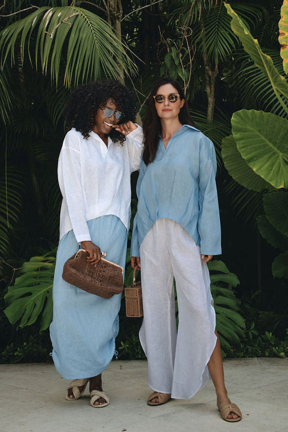 Two women posing in front of a lush tropical garden backdrop. The woman on the left wears a white long-sleeve top and a 100% linen light blue maxi skirt, holding a brown leather handbag.