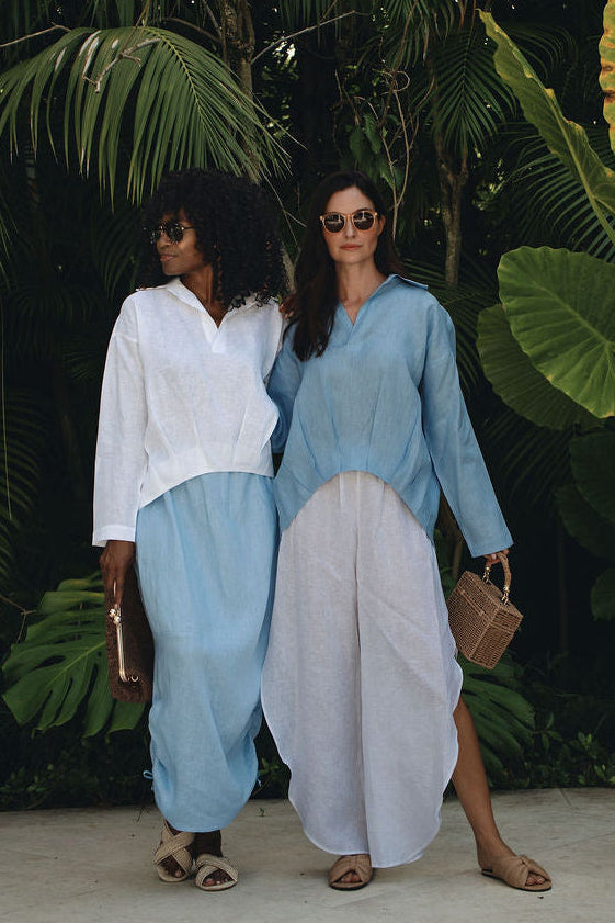 Two women posing in front of lush tropical green foliage. One is wearing a white long-sleeve top and light blue wide-leg pants, while the other wears a light blue top and a white skirt.