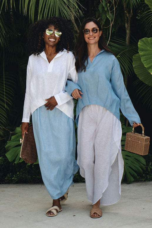 Two women walking hand in hand outdoors in front of lush tropical green foliage. The woman on the left wears a white long-sleeve linen top and light blue wide-leg pants, while the woman on the right wears a light blue linen long-sleeve top and linen white wide-leg pants.