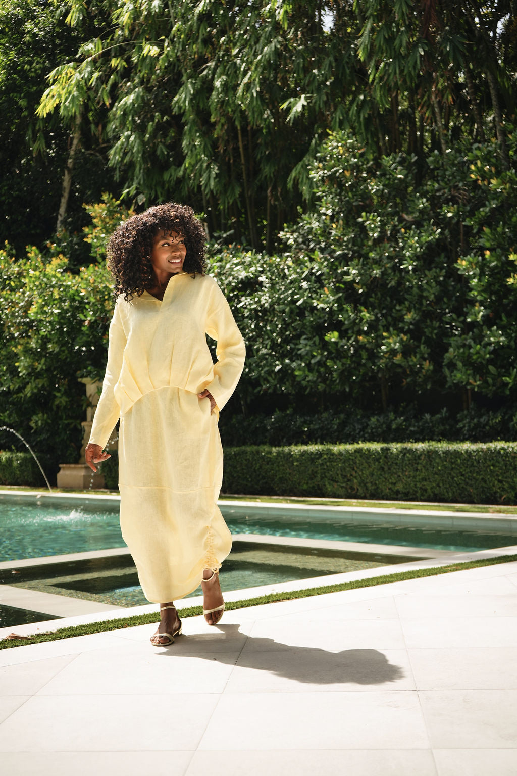 A woman with an afro walking outdoors near a pool, wearing a loose, long-sleeve yellow linen shirt with a linen skirt with pockets and brown sandals.