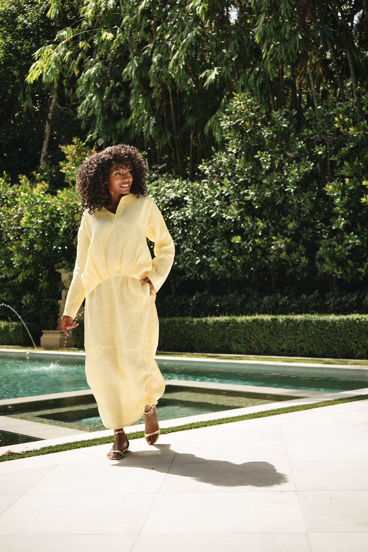 A woman with an afro walking outdoors near a pool, wearing a loose, long-sleeve yellow linen shirt with a linen skirt with pockets and brown sandals.