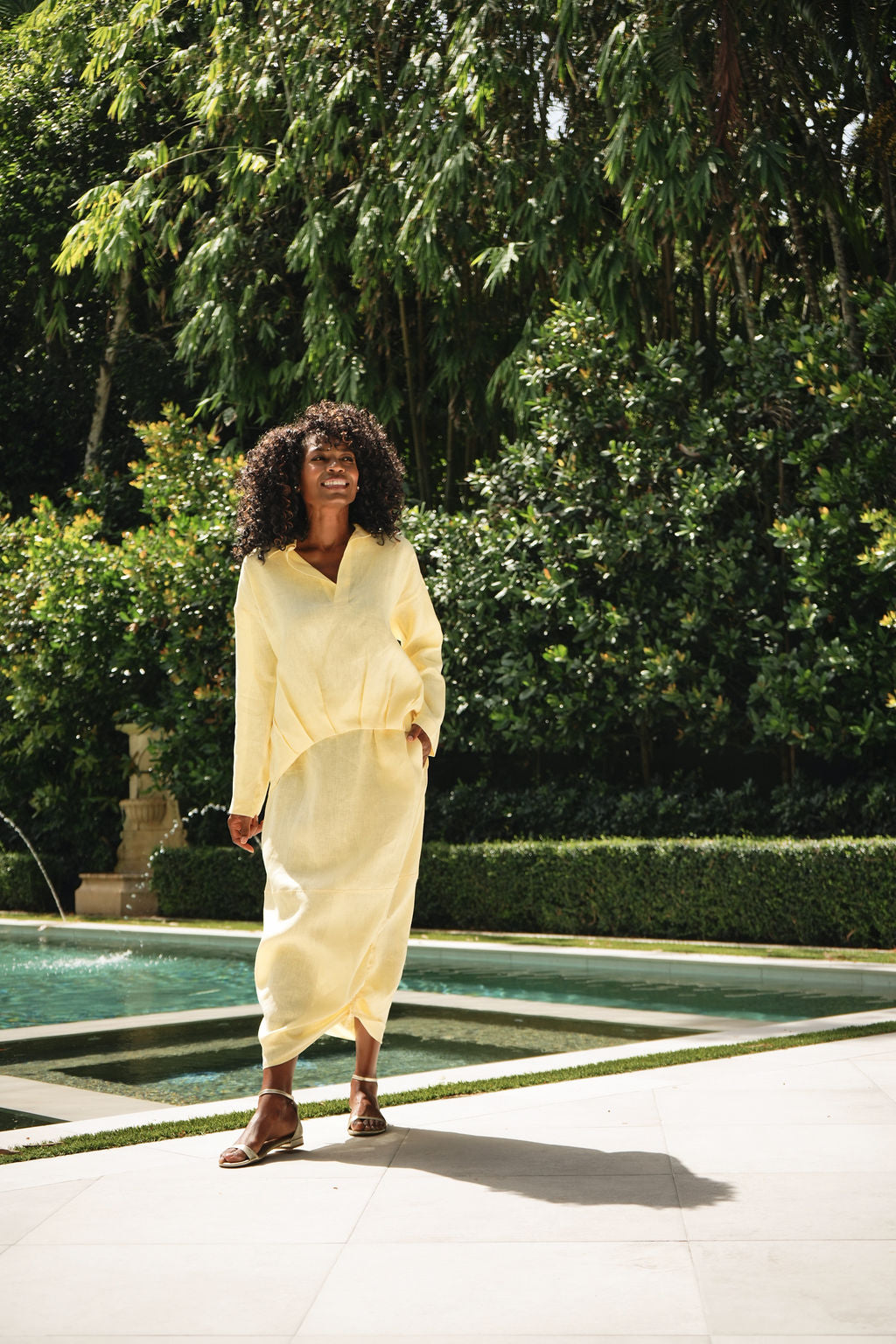 A woman wearing a yellow linen long-sleeve shirt walks outdoors in Coral Gables beside a swimming pool in a lush garden setting.