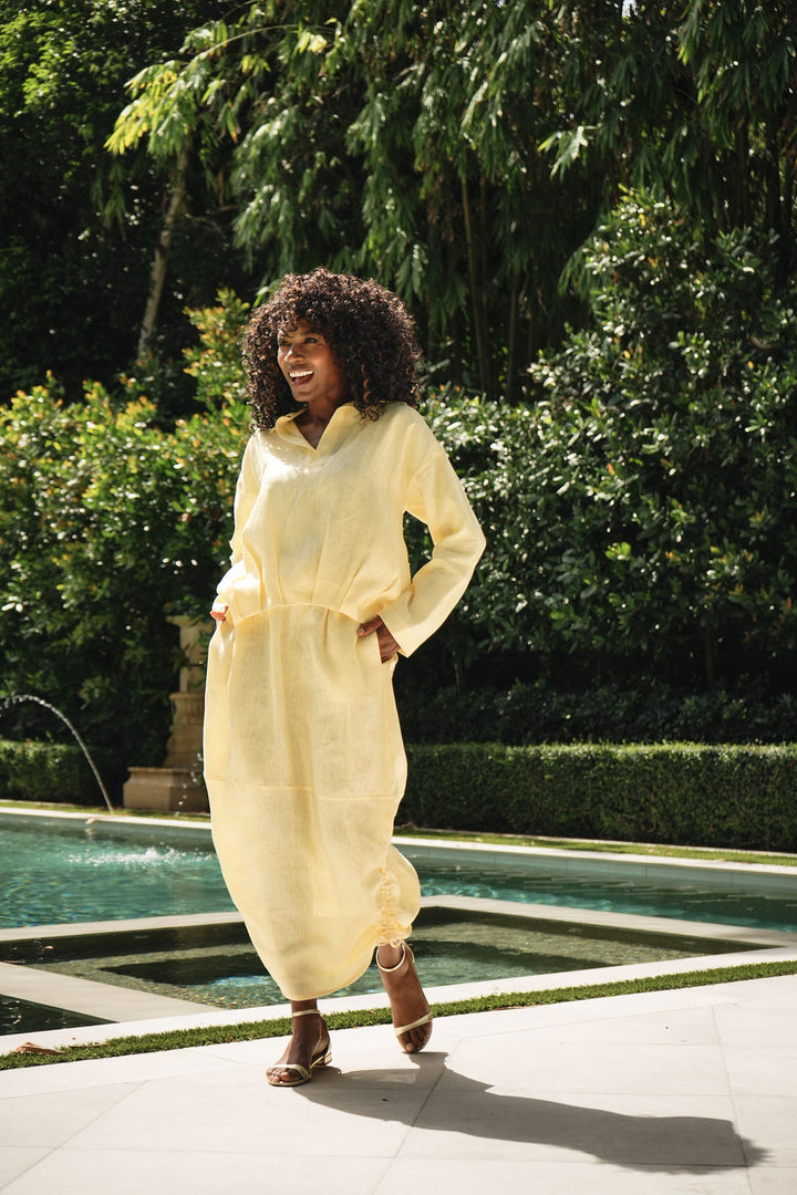 A woman wearing a yellow linen long-sleeve shirt and skirt with pockets walks beside a swimming pool in a lush garden setting.