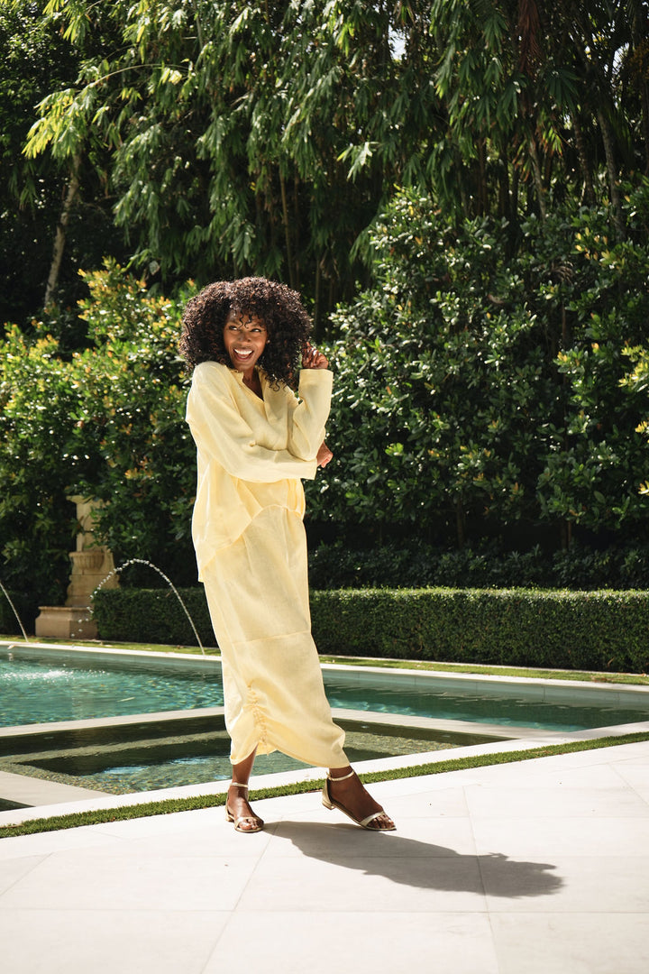 A woman with curly dark hair stands by a swimming pool in a lush garden, wearing a loose, long-sleeve yellow linen shirt and matching linen skirt.